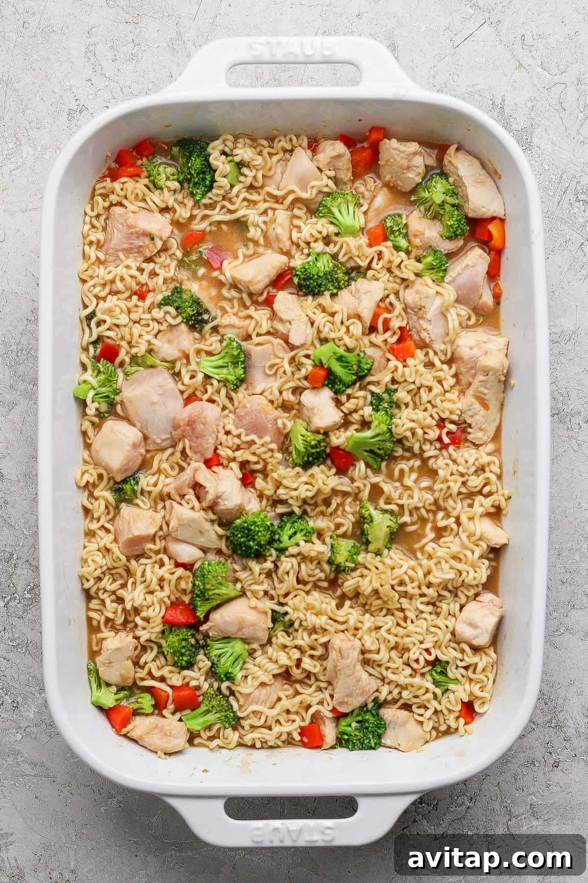 A white baking dish filled with cooked ramen noodles, pieces of chicken, broccoli florets, and red bell pepper in a light broth.