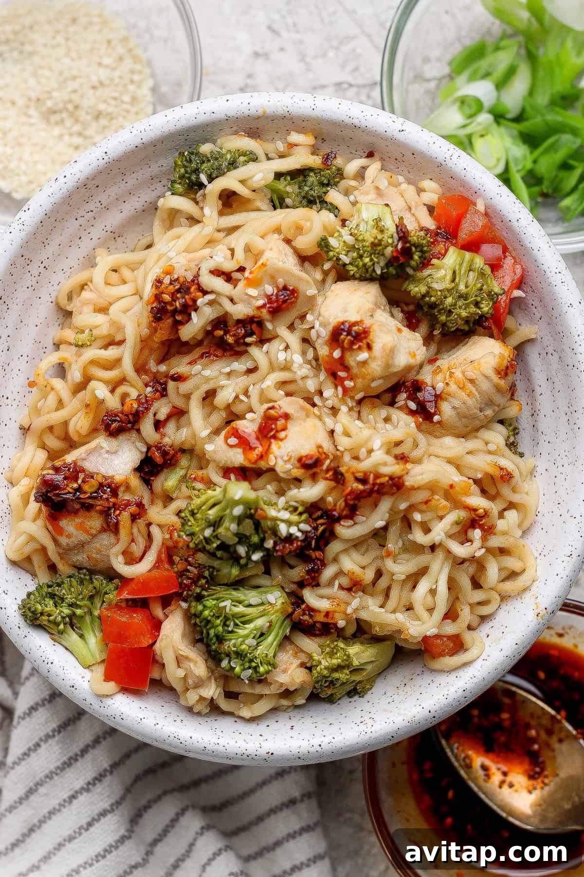 A bowl of ramen noodles mixed with chicken, broccoli, and red bell peppers, topped with chili oil and sesame seeds, with green onions and sauces on the side.
