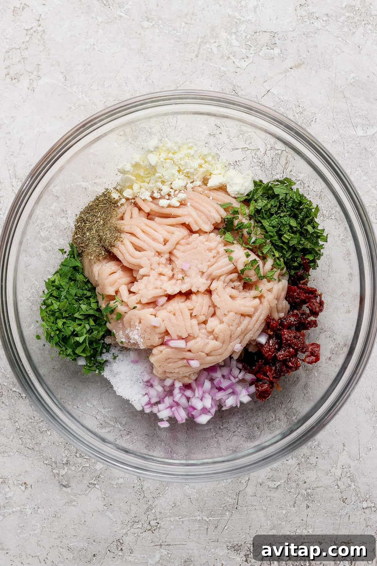 A glass bowl containing ground meat, chopped red onion, herbs, minced garlic, sun-dried tomatoes, salt, pepper, and seasonings arranged separately.