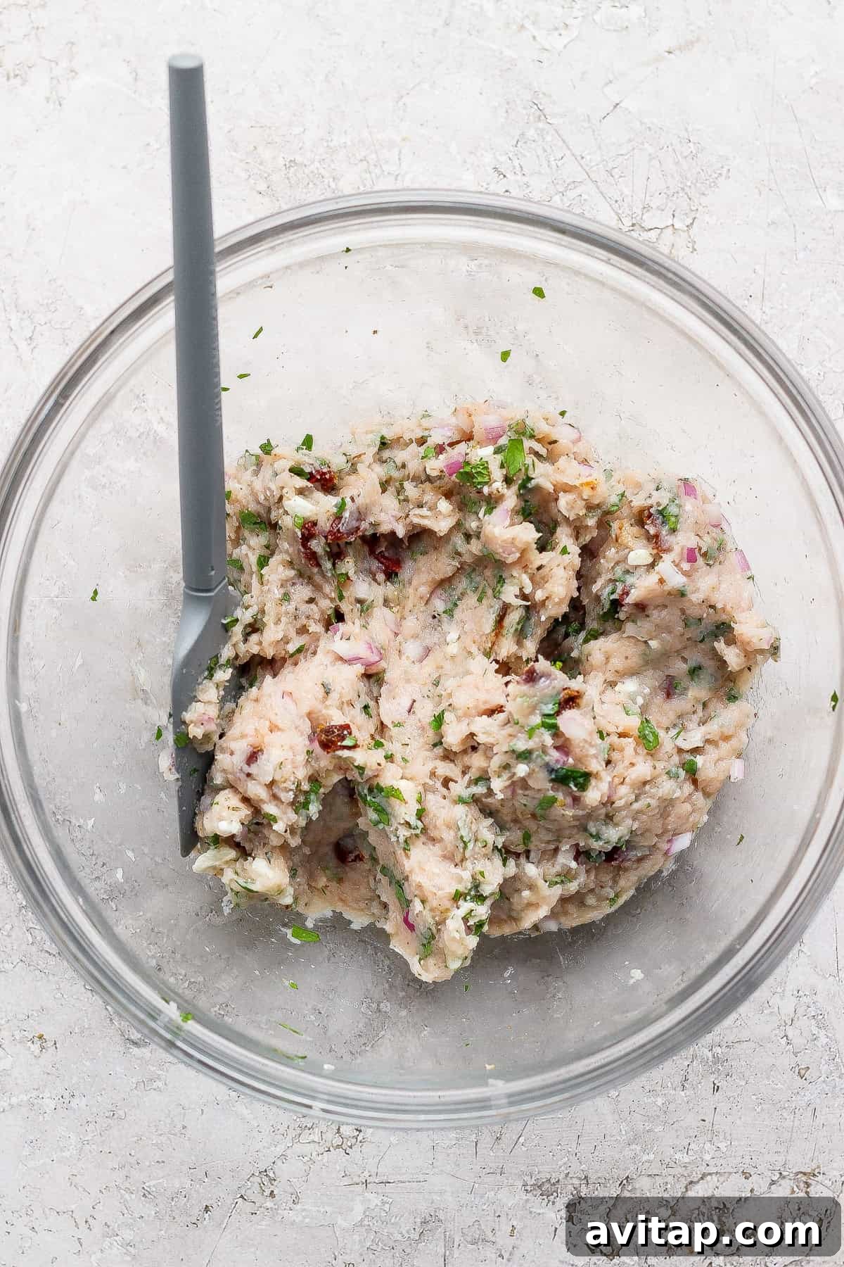 A glass bowl with a gray spoon contains a mixture of mashed white beans, chopped herbs, red onion, and sun-dried tomatoes on a light-colored surface.