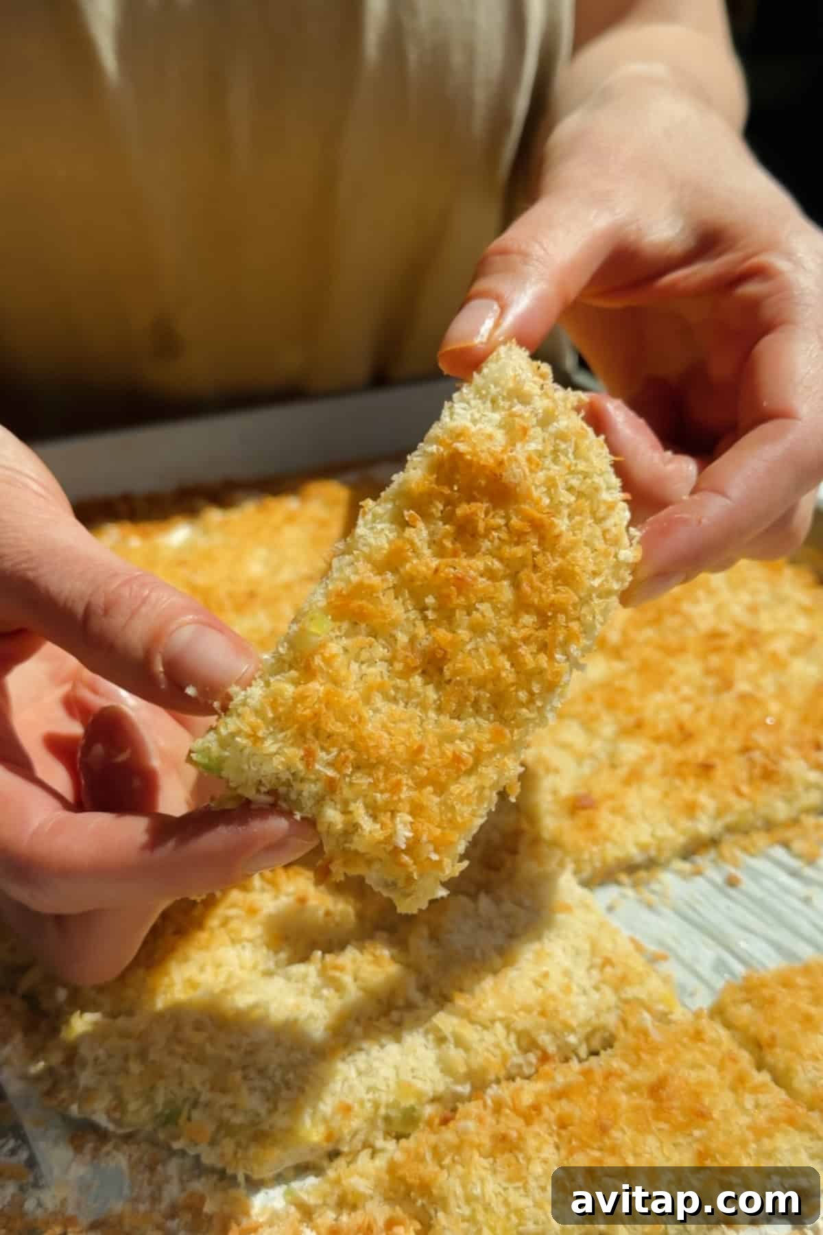 Buffalo Chicken Protein Bars — Crispy Baked Snackable Bars 3 Holding a golden crispy chicken protein bar with panko coating above a tray of baked bars