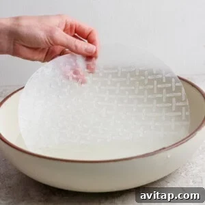 A hand dips a round sheet of rice paper into a shallow bowl of water on a light-colored surface.