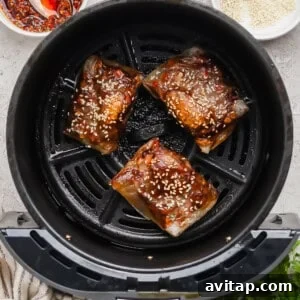 Three pieces of marinated fish with sesame seeds in an air fryer basket, with a bowl of chili sauce and a bowl of sesame seeds nearby.