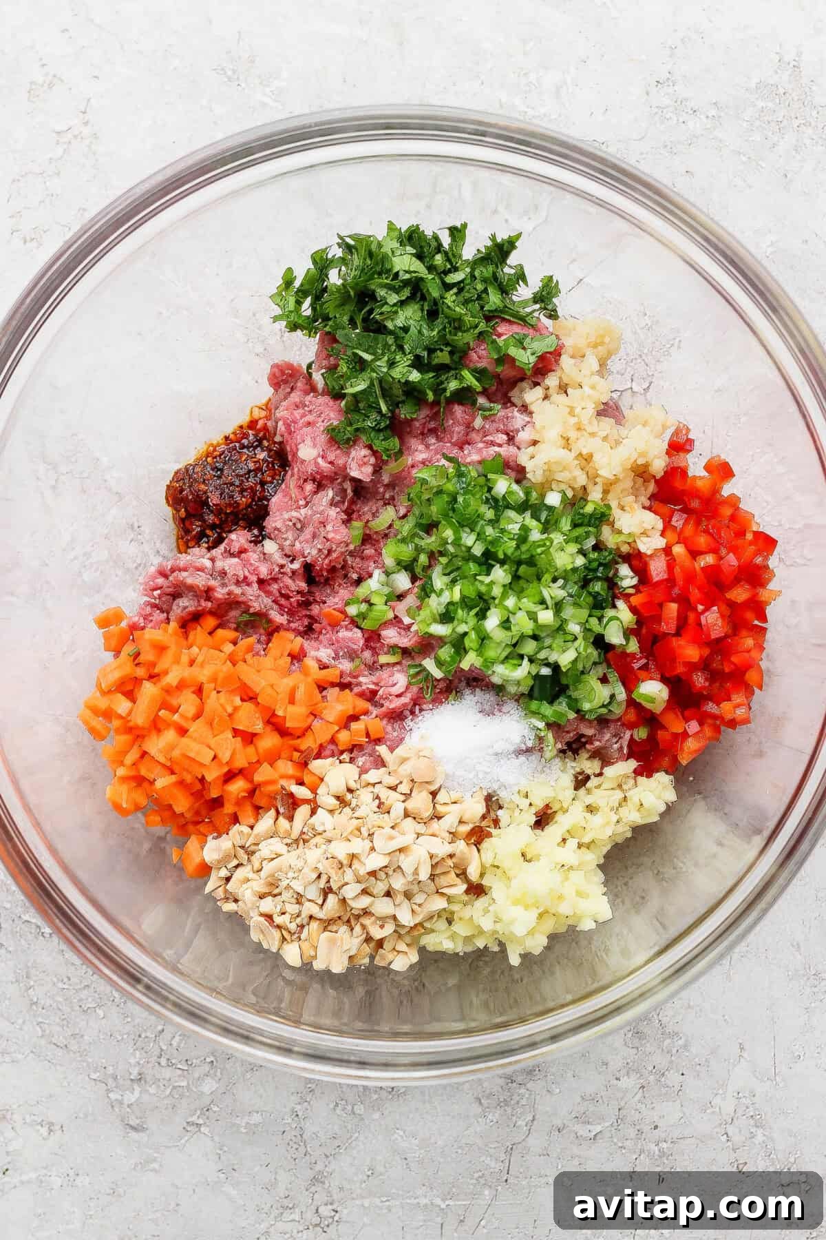A glass bowl containing ground meat, chopped carrots, bell peppers, mushrooms, parsley, ginger, garlic, green onions, and seasonings arranged in separate piles.