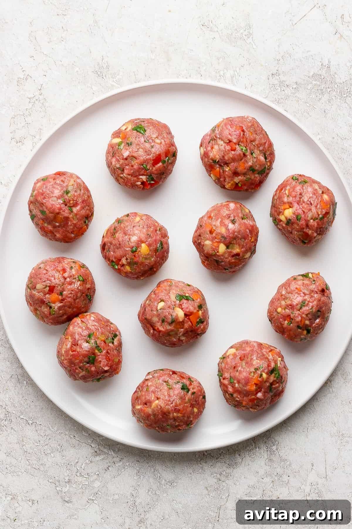 A white plate with twelve uncooked meatballs, mixed with herbs and small vegetable pieces, on a light textured surface.