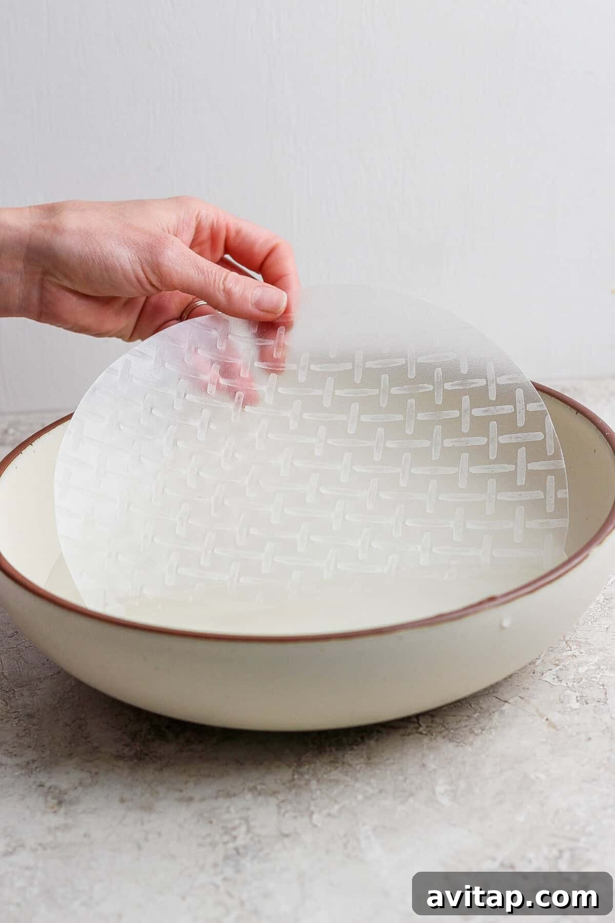 A hand dips a round sheet of rice paper into a shallow bowl of water on a light-colored surface.