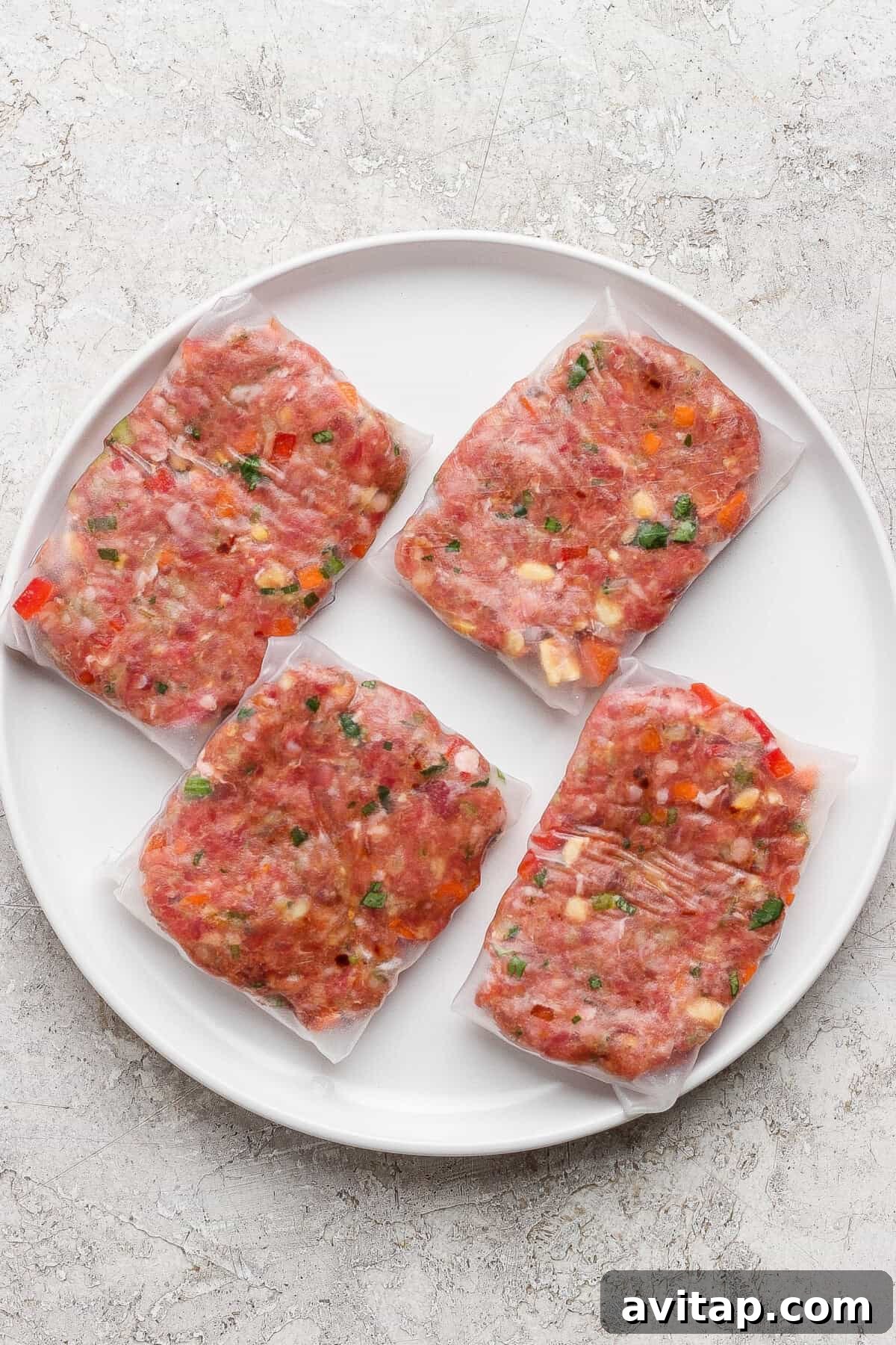 Four vacuum-sealed portions of seasoned raw ground meat with visible herbs and vegetables, arranged on a round white plate.