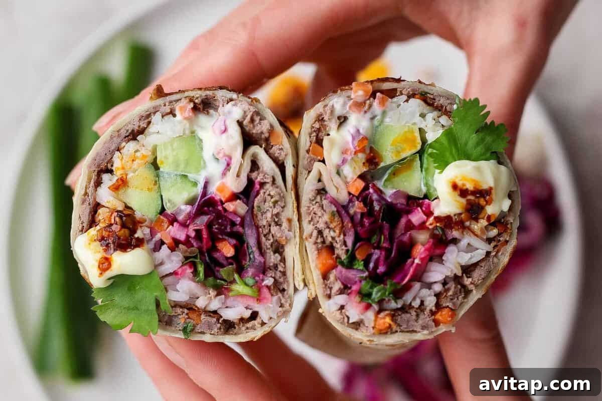 A close-up of hands holding a cut burrito filled with rice, ground meat, cucumber, egg, shredded vegetables, cilantro, and sauce, with a plate of food in the background.