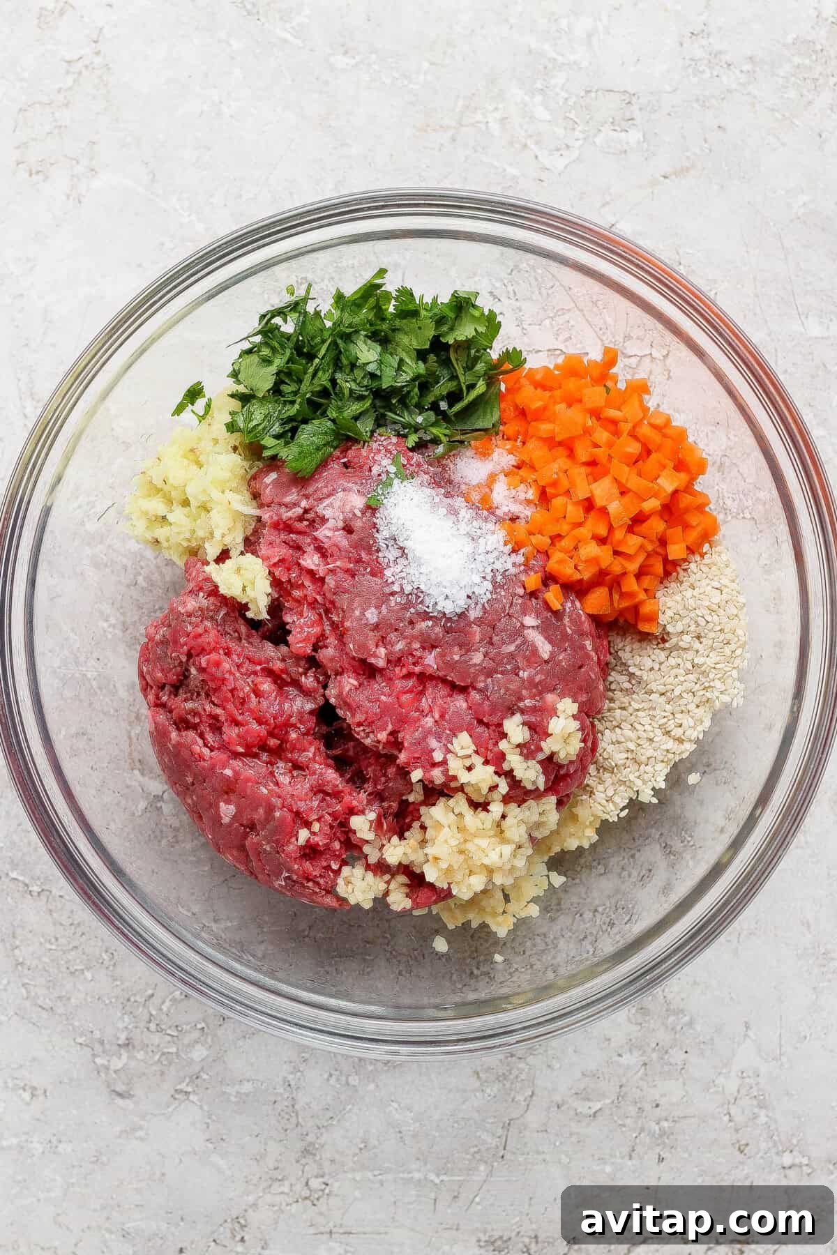 A glass bowl containing ground beef, chopped parsley, diced carrots, minced garlic, breadcrumbs, and salt on a light countertop.