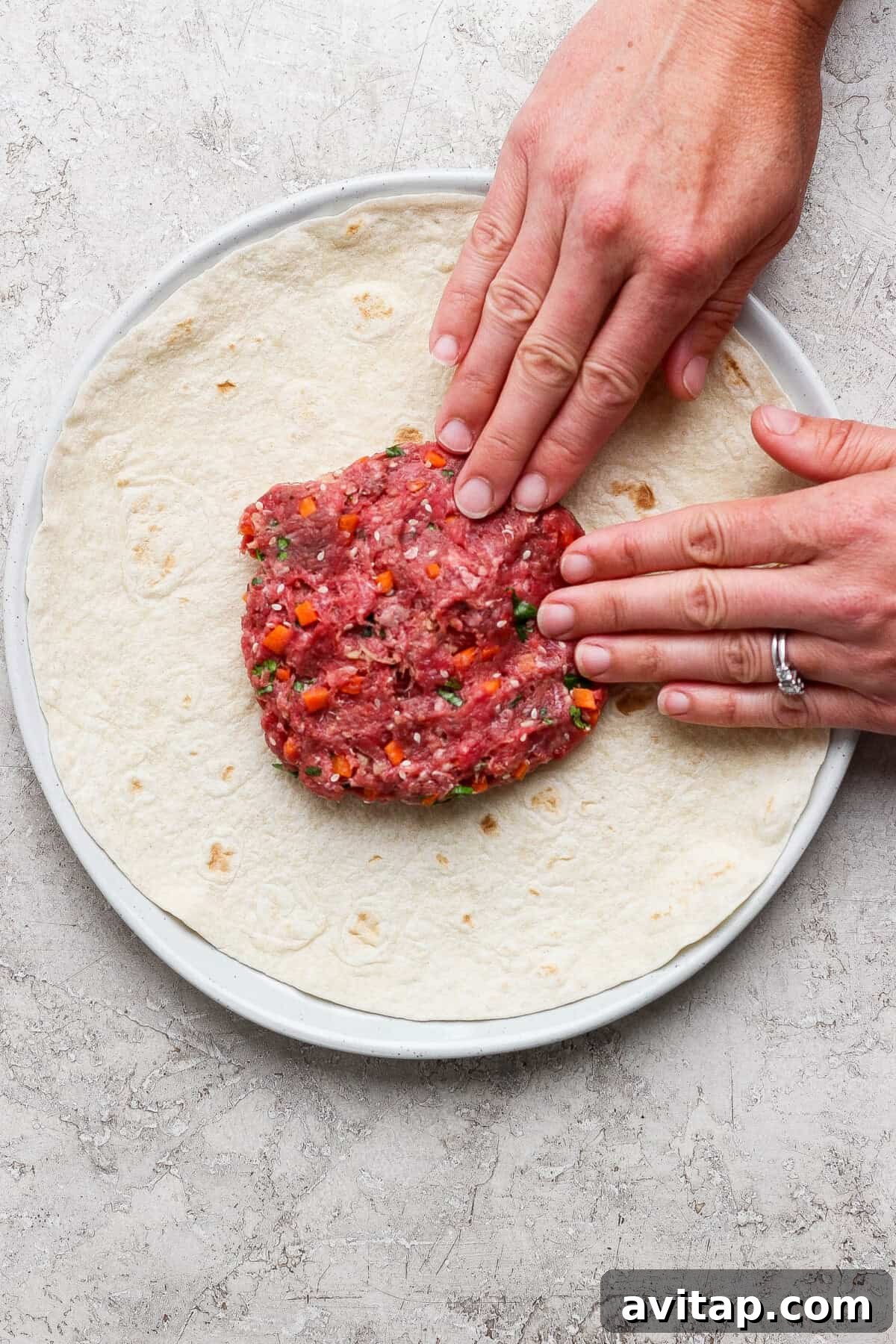Hands pressing raw ground meat mixture with chopped vegetables onto a large tortilla on a plate.