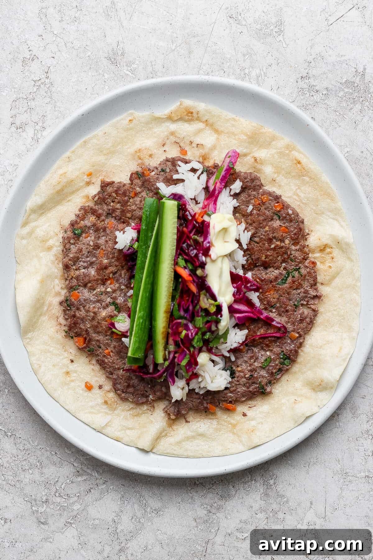 A plate with a flatbread topped with ground meat, rice, red cabbage slaw, cucumber sticks, and a drizzle of white sauce.