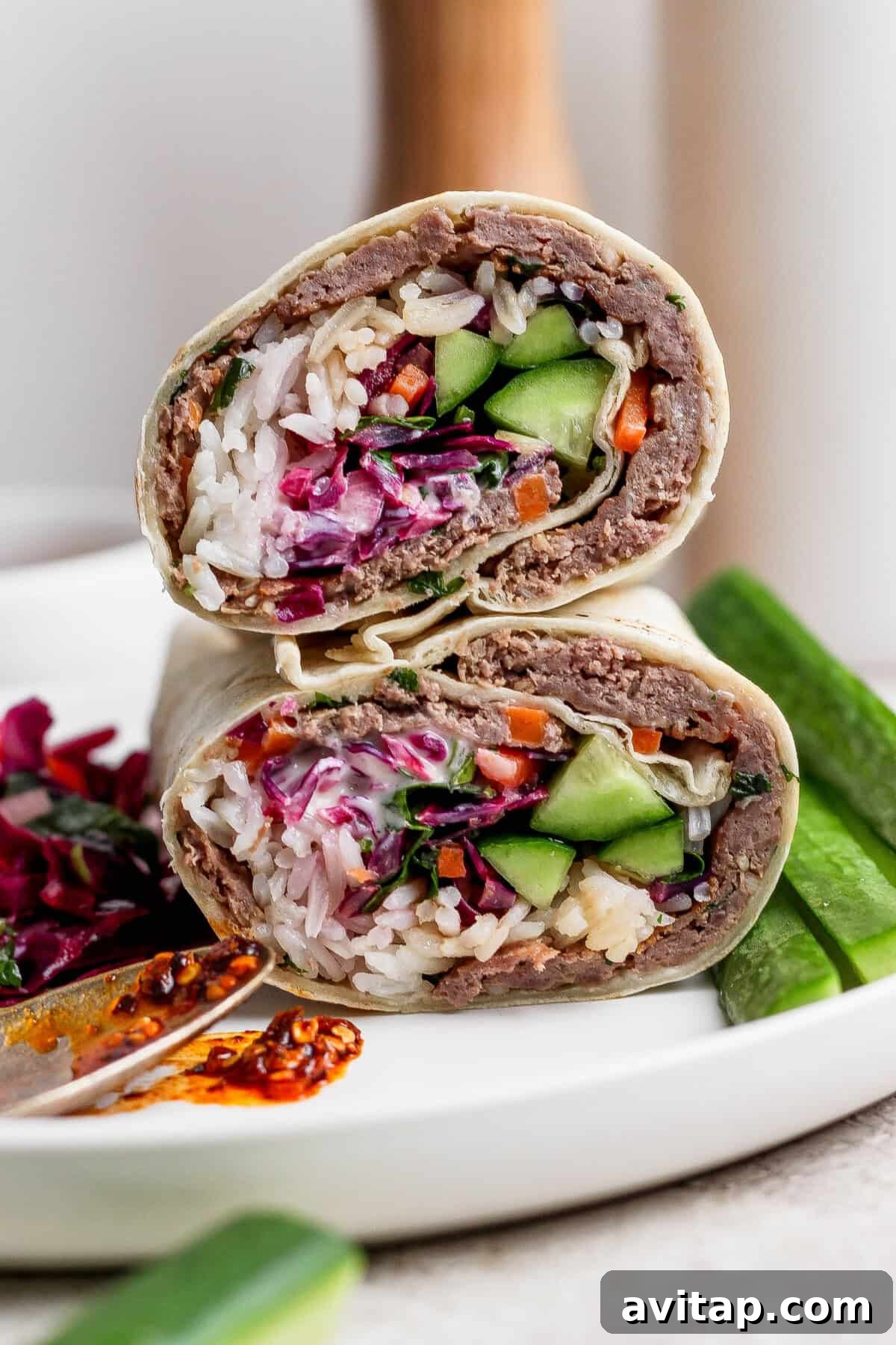 A close-up of a halved wrap filled with rice, vegetables, and a ground meat mixture, served on a white plate with cucumber slices and a side of spicy sauce.