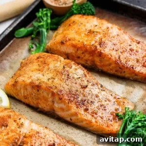 Three golden broiled salmon fillets on a parchment-lined baking sheet with broccolini and lemon.