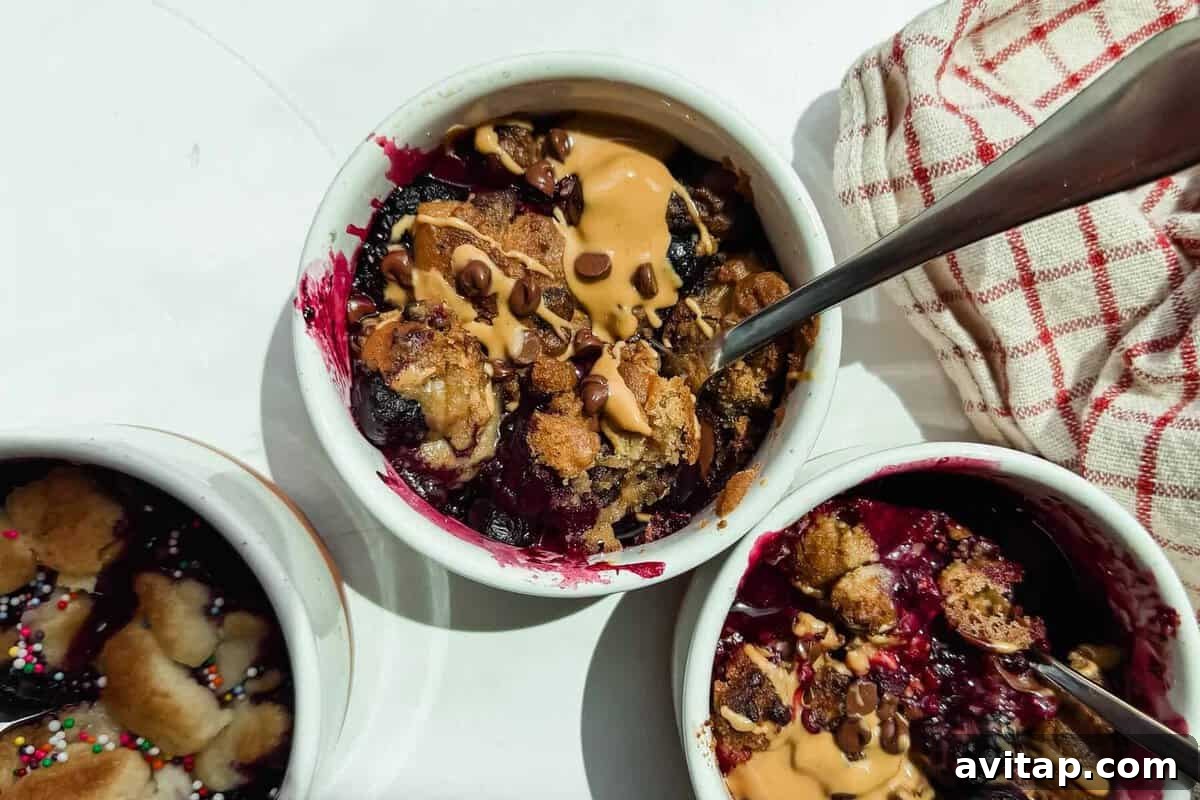 Three small white ramekins filled with baked berry dessert, topped with peanut butter and chocolate chips, with metal spoons and a red-striped cloth nearby.
