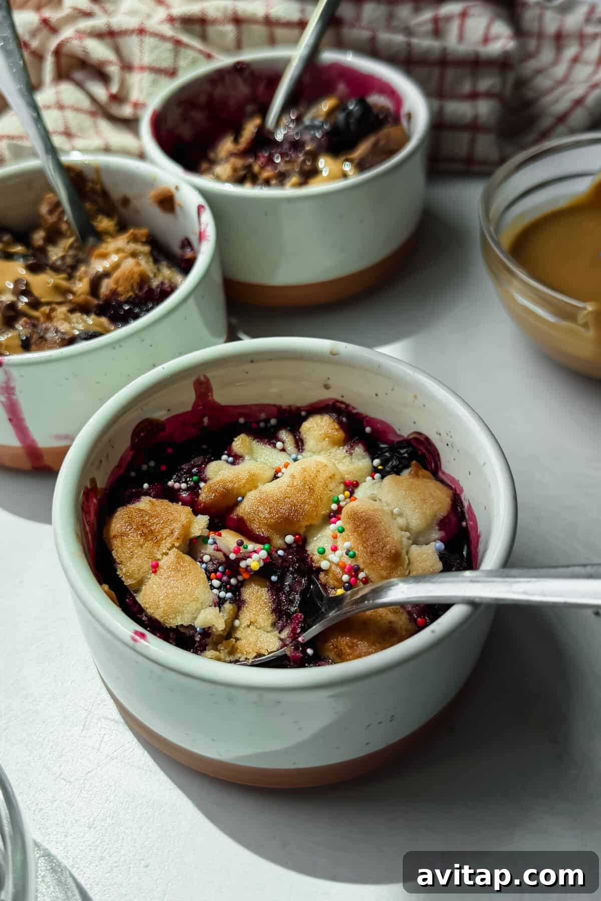 Three ceramic bowls of berry crumble topped with sprinkles and shortbread pieces, each with a spoon, sit on a light surface next to a bowl of caramel sauce.
