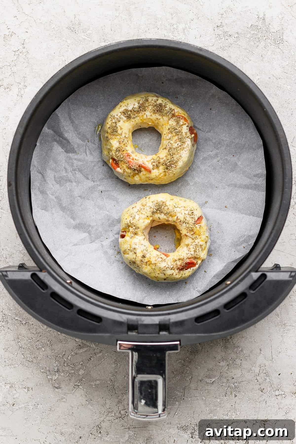 Two uncooked pizza protein bagels with seasoning in an air fryer basket