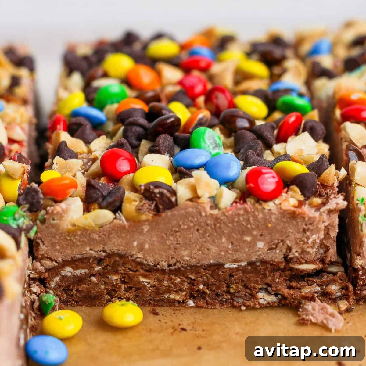 Close-up of dessert bars topped with colorful candy-coated chocolates, chocolate chips, and chopped nuts on a brown surface.