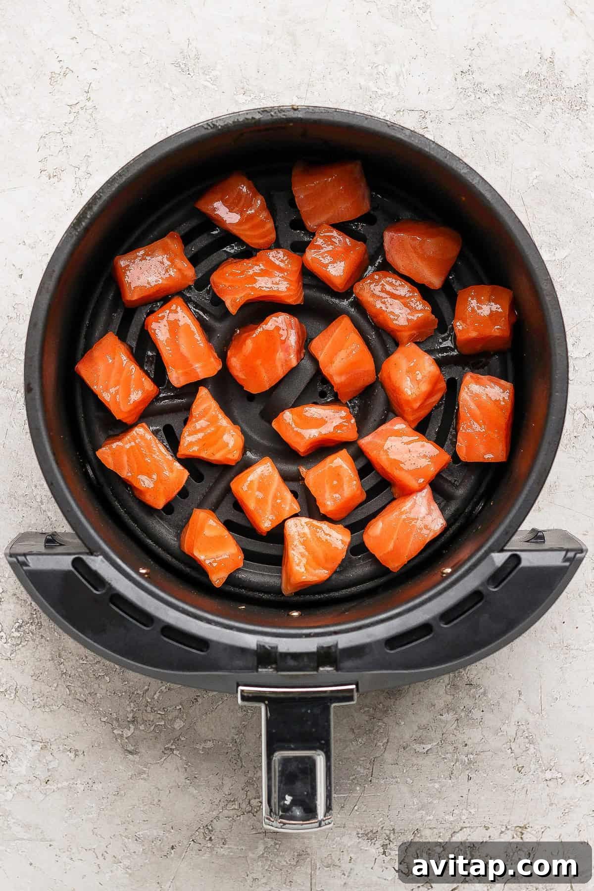 Raw salmon cubes are arranged in a single layer inside the basket of an air fryer on a light-colored surface.