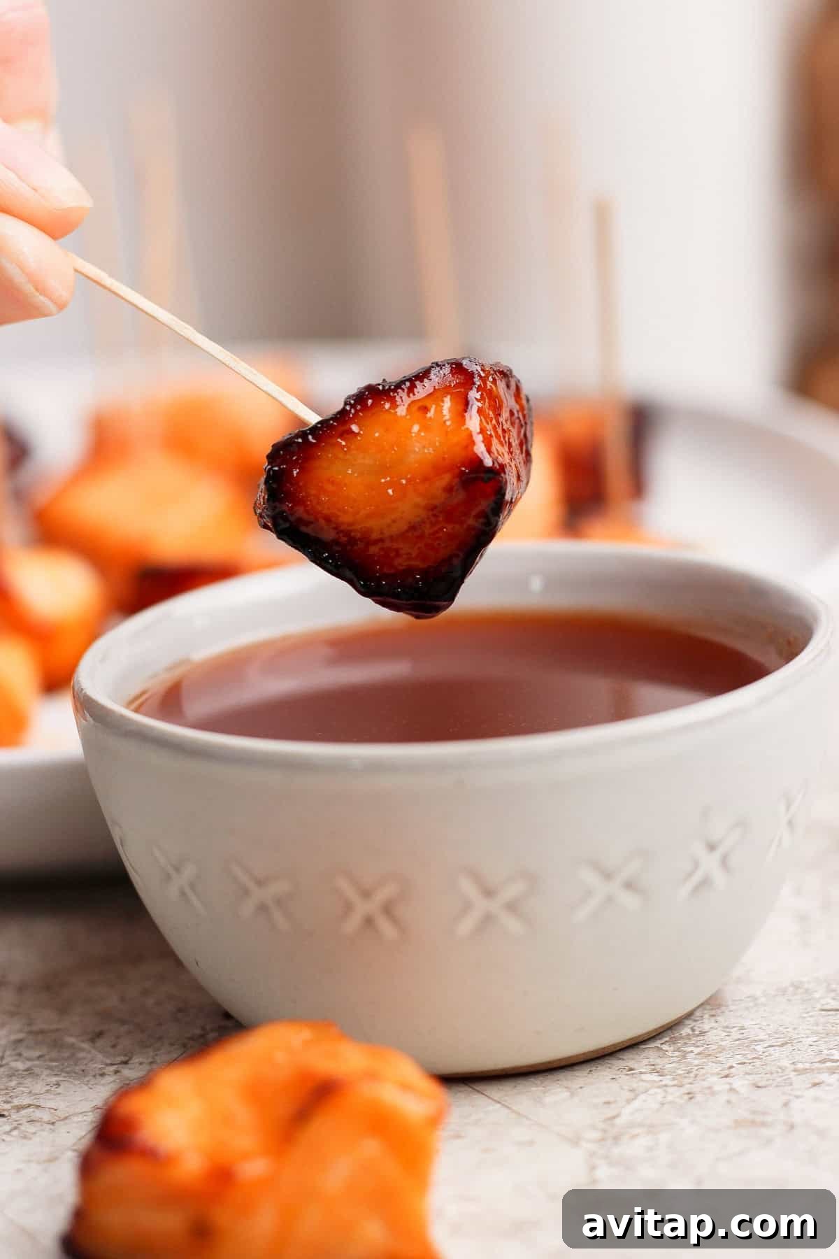 A hand holds a skewer with a glazed, roasted bite-sized food piece above a bowl of dipping sauce; more skewered pieces are seen in the background.