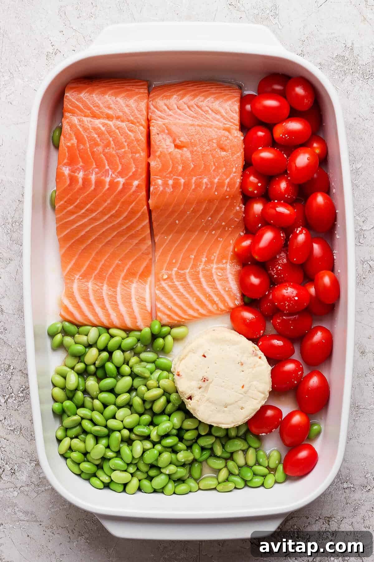 Raw salmon fillets, cherry tomatoes, edamame, and Boursin cheese assembled in a white casserole dish before baking