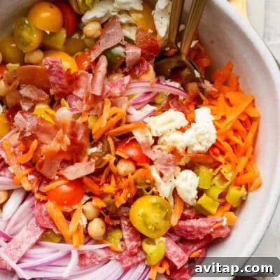 A salad bowl with shredded carrots, sliced red onions, cherry tomatoes, chickpeas, feta cheese, chopped bacon, and salami.