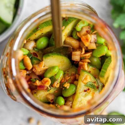 Top view of a jar filled with sliced cucumbers, edamame, chopped nuts, red onion, and spices.