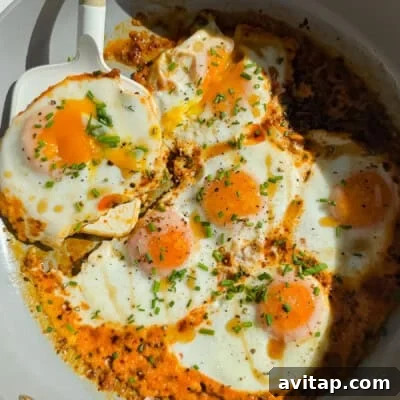 Six sunny-side up eggs cooking in a skillet with herbs and spices, one being lifted by a white spatula.