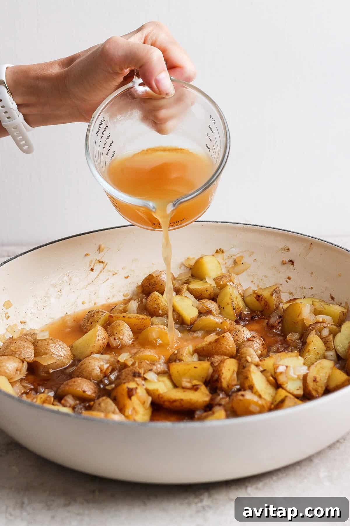 Apple cider being poured into a skillet of browned baby potatoes, onion, and garlic.