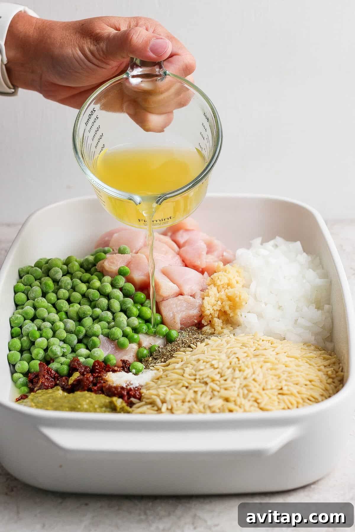 Chicken broth being poured over raw chicken, dry orzo, peas, pesto, sun-dried tomatoes, onion, garlic, and seasonings in a casserole dish.