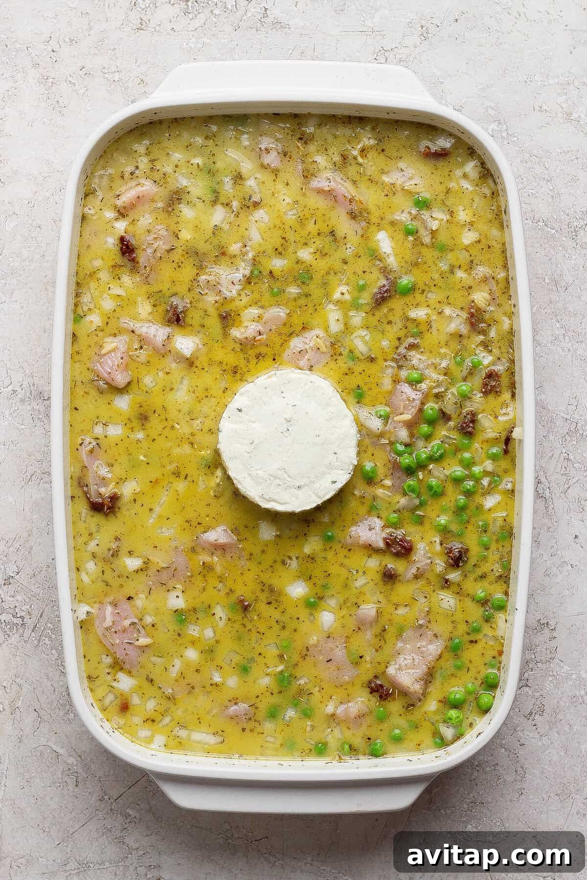 Unbaked chicken pesto orzo mixture in a white casserole dish with a whole puck of Boursin in the center.