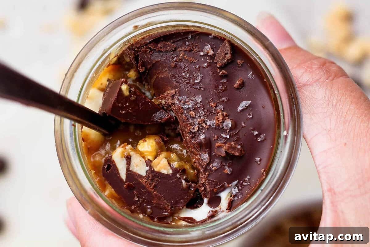 Spoon breaking through the chocolate shell on an individual Snickers cheesecake cup to reveal peanut butter and cottage cheese layers.